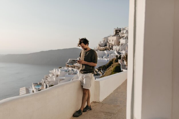 Encantador hombre en pantalones cortos blancos camiseta verde oscuro chats en teléfono fuera Morena chico feliz posa de fondo de la ciudad y el mar