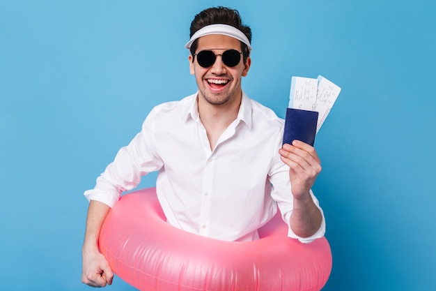 Encantador joven en camisa blanca clásica, gafas de sol y gorra sonríe con alegría y sostiene documentos, círculo inflable.