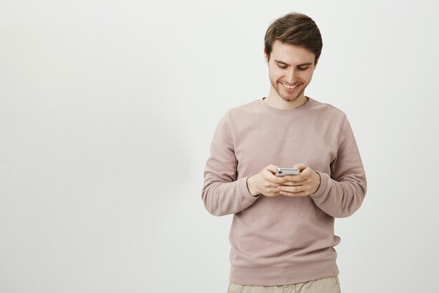 Encantador joven con linda sonrisa y cerdas sosteniendo teléfono inteligente y mensajería