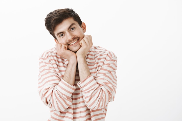 Encantador y lindo novio con bigote, apoyando la cabeza en las manos y sonriendo amigablemente, mirando con admiración, escuchando atentamente el discurso del atractivo conferenciante, de pie sobre una pared gris