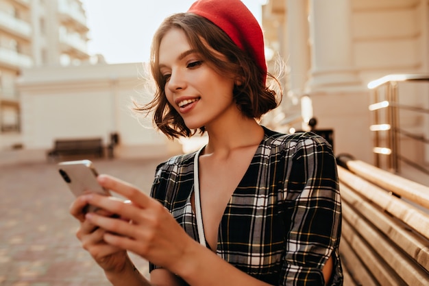 Encantador mensaje de texto elegante chica en día de otoño. Debonair mujer de pelo corto en boina roja de pie en la calle con teléfono.