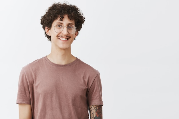 Foto gratuita encantador, motivado, guapo, chico de pelo rizado con bigote y brazo tatuado con anteojos y una camiseta de moda, sonriendo con alegría y sentimiento amistoso cumplido subiendo a los sueños