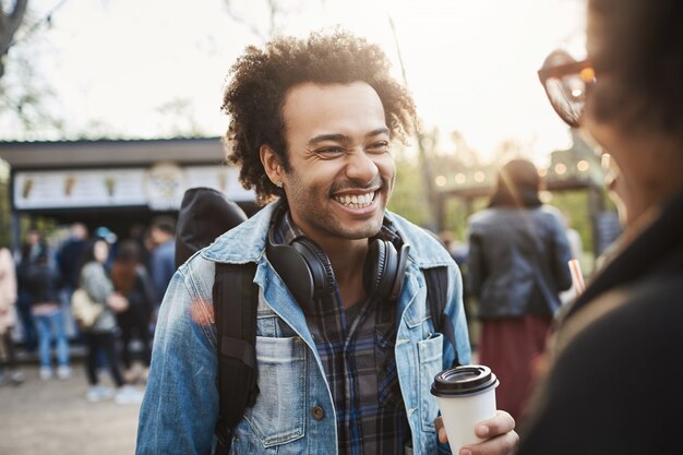 Encantador novio feliz con peinado afro sonriendo y riendo mientras habla con su novia y toma café en el parque.