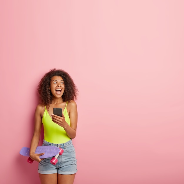 Encantadora adolescente llena de alegría con tupido cabello afro, sostiene el teléfono móvil