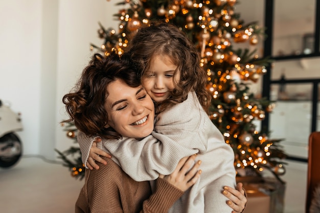 Encantadora adorable mujer caucásica con curvas abrazándose con su pequeña hija y celebrando la Navidad y el Año Nuevo