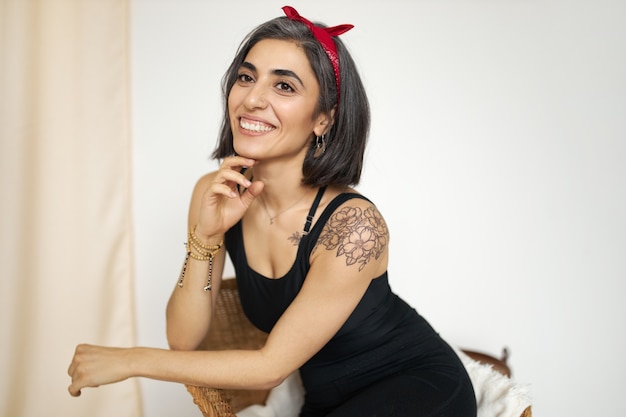 Encantadora y amigable joven mujer de raza mixta con figura ajustada, tatuaje y linda sonrisa posando en el interior