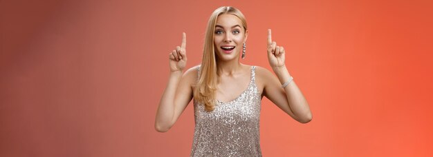 Encantadora y asombrada mujer europea rubia en un fabuloso vestido plateado y brillante levanta las manos apuntando hacia arriba divertida