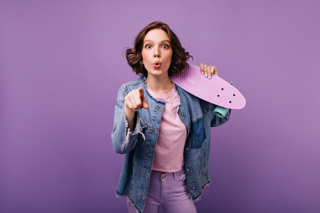 Encantadora chica activa en chaqueta de mezclilla que expresa emociones de sorpresa. Foto interior de mujer morena bastante rizada con patineta.