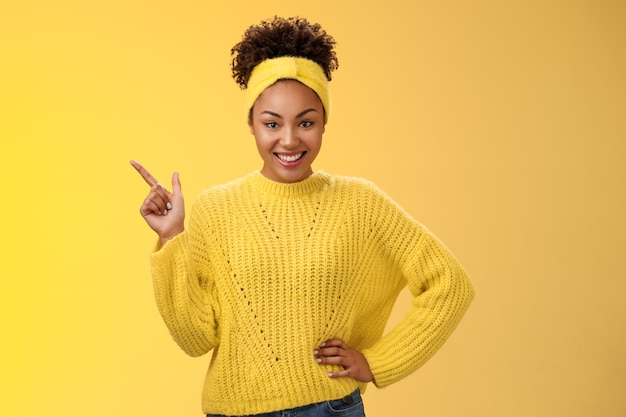 Encantadora chica afroamericana con estilo descarada en suéter apuntando en la esquina superior izquierda de pie con una sonrisa atrevida y segura con fondo amarillo que promueve el merchandising que muestra la mejor opción, mira la cámara divertida.