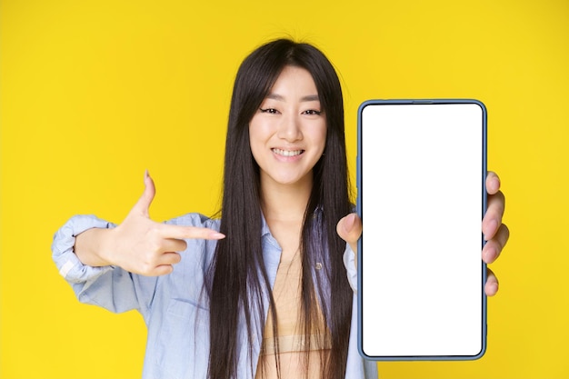 Encantadora chica asiática con teléfono inteligente en la mano señalando con el dedo en la pantalla blanca Gran oferta Colocación de productos publicitarios de aplicaciones móviles