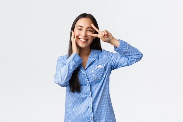 Encantadora chica asiática tonta en pijama azul mostrando el signo de la paz y tocando la mejilla kawaii, sonrojándose y sonriendo, satisfecha con una piel limpia perfecta, usa productos para el cuidado de la piel, fondo blanco.