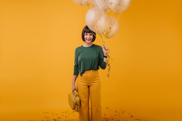 Encantadora chica con bolso amarillo posando con una sonrisa después de la fiesta. Foto interior de hermosa dama morena con globos.