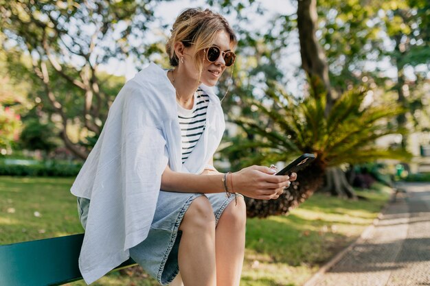 Encantadora chica bonita con cabello ondulado recogido con gafas de sol, camisa azul y camiseta a rayas sentada en el banco en el jardín de la ciudad a la luz del sol y desplazando el teléfono inteligente con una sonrisa