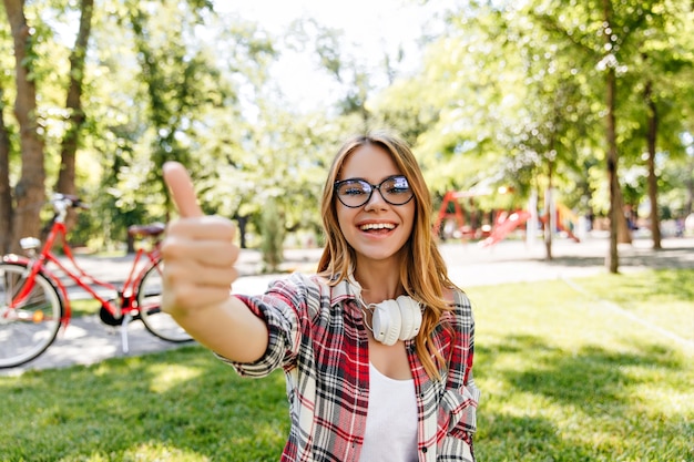 Encantadora chica en camisa a cuadros posando en la naturaleza. Señora caucásica alegre en vasos pasar tiempo en el parque.