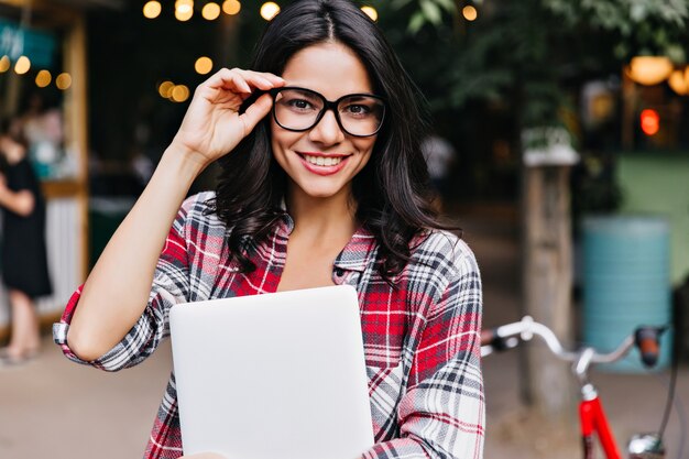 Encantadora chica caucásica en gafas posando después de clases. Foto al aire libre de estudiante alegre con portátil.