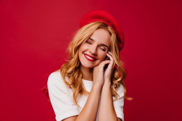 Encantadora chica caucásica con peinado ondulado riendo en pared roja. alegre modelo femenino francés en boina.