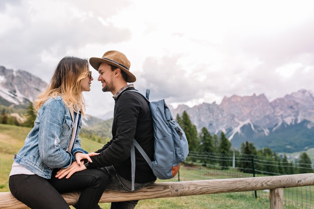 Encantadora chica en chaqueta de mezclilla vintage relajante con su novio pasar tiempo al aire libre con montañas