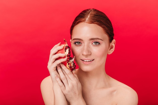 Encantadora chica desnuda sosteniendo granate y sonriente Foto de estudio de mujer atractiva de buen humor posando con granada sobre fondo rojo