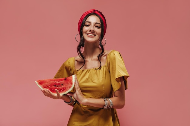 Encantadora chica elegante con cabello oscuro ondulado en accesorios elegantes y vestido amarillo sonriendo y sosteniendo sandía sobre fondo rosa