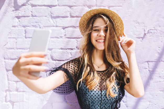 Encantadora chica con elegante collar haciendo selfie delante de la vieja pared blanca