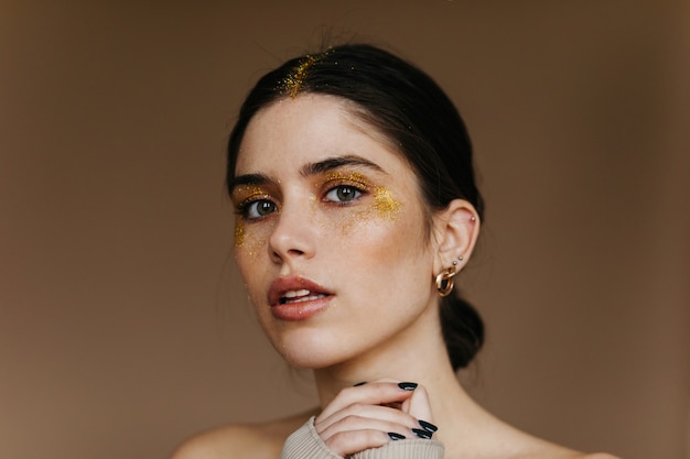 Encantadora chica con elegante maquillaje dorado. Foto de primer plano interior de dama entusiasta con cabello negro.