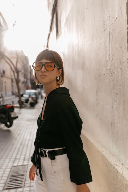 Encantadora chica elegante con peinado corto con blusa negra y gafas amarillas de moda posando en la cámara en las noches de verano a la luz del sol