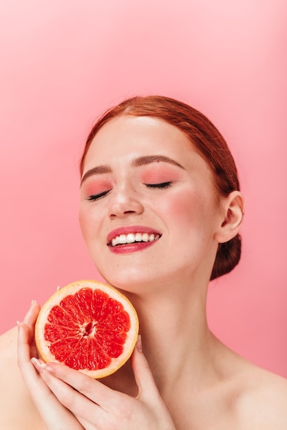 Encantadora chica europea con pomelo jugoso. Foto de estudio de mujer riendo feliz con cítricos posando con los ojos cerrados sobre fondo rosa.