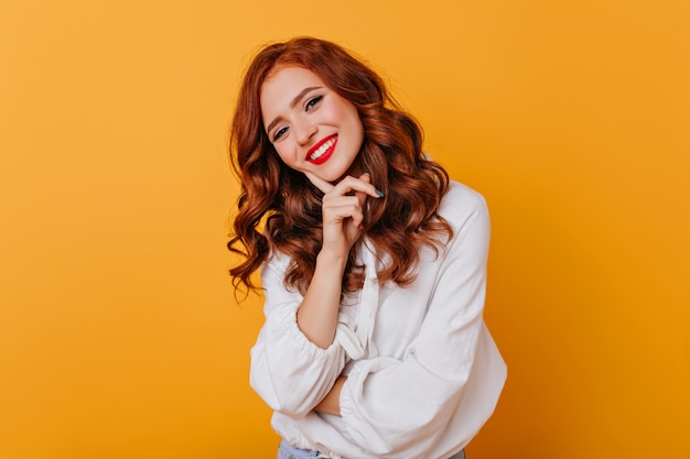 Encantadora chica europea viste blusa blanca posando. Foto interior de elegante mujer pelirroja sonriendo en pared brillante.