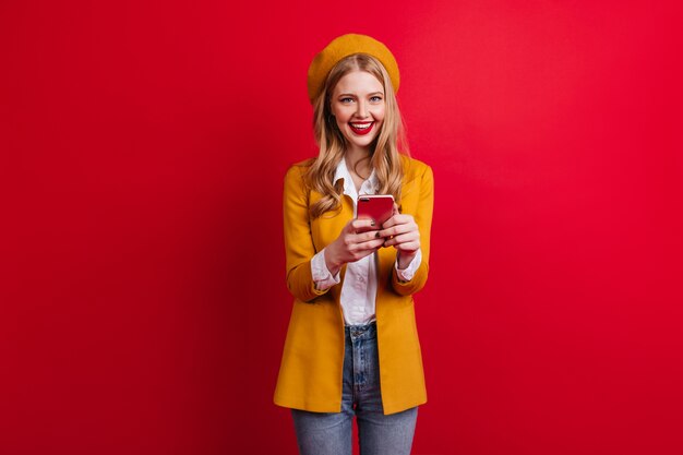 Encantadora chica francesa con teléfono en la pared roja. riendo mujer rubia en chaqueta amarilla con smartphone.