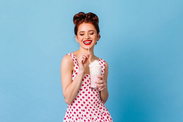 Encantadora chica de jengibre bebiendo batido. Disparo de estudio de dama feliz en vestido de lunares disfrutando de bebidas en el espacio azul.