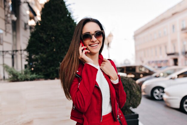 Encantadora chica lleva grandes gafas de sol marrones hablando por teléfono en la mañana