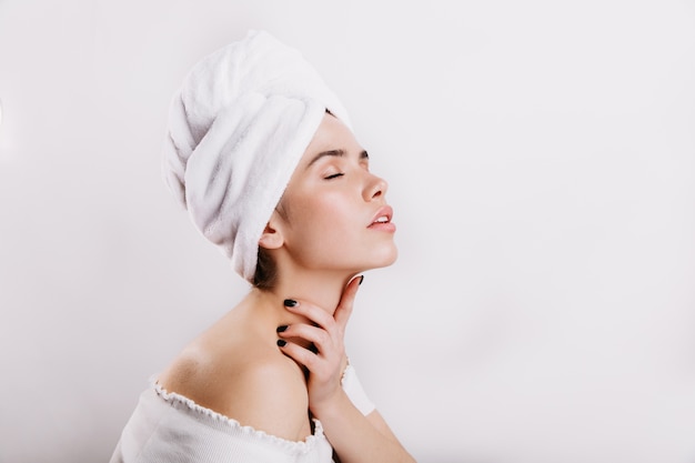 Encantadora chica sin maquillaje masajea suavemente su cuello. Mujer con una piel perfecta posando en la pared blanca.