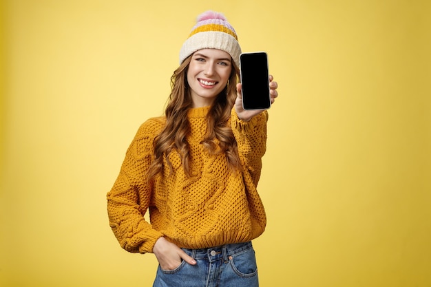 Encantadora chica de moda sonriente saliente extiende el brazo mostrando tu nuevo teléfono inteligente, muestra sonriendo satisfecho a un amigo consultor qué filtro puso usando la aplicación editar foto teléfono móvil, fondo amarillo