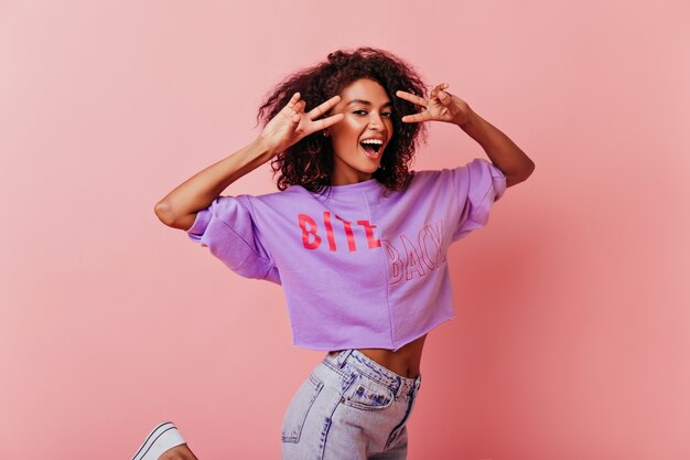 Encantadora chica morena divertida posando con el signo de la paz. Retrato de estudio de mujer alegre aislada en rosa.