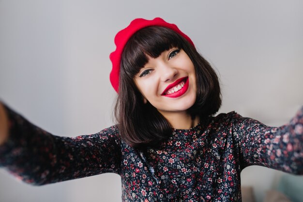 Encantadora chica morena con maquillaje brillante sonriendo ampliamente mientras hace selfie en su habitación. Retrato de primer plano de adorable joven morena vestida con lindo traje vintage y boina francesa roja