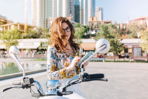 Encantadora chica morena posando juguetonamente con el teléfono en las manos de pie en la plaza con rascacielos