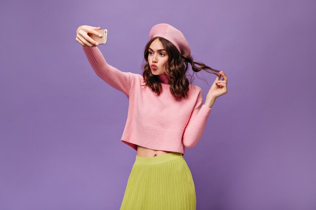 Encantadora chica morena toca el cabello, sostiene el teléfono inteligente y toma selfie