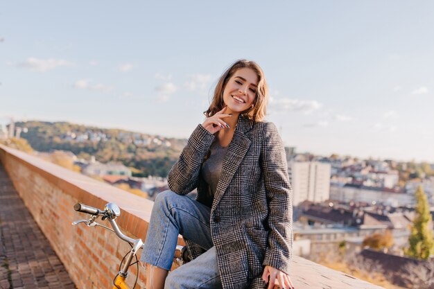 Encantadora chica morena viste chaqueta elegante y jeans posando sobre fondo de ciudad hermosa