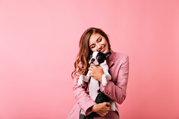 Encantadora chica con peinado largo abrazando bulldog francés con los ojos cerrados. Retrato interior de modelo femenino de jengibre despreocupado con lindo perrito y riendo.