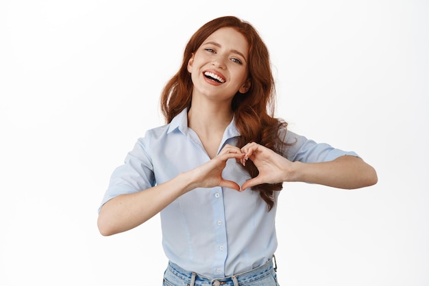 Encantadora chica pelirroja muestra el signo del corazón y sonríe a la cámara te amo gesto estado de ánimo romántico de pie en la blusa contra el fondo blanco.