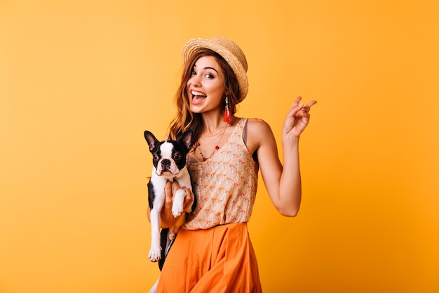 Encantadora chica pelirroja con sombrero vintage con bulldog francés. Retrato interior de mujer blanca asombrada posando en amarillo con cachorro.