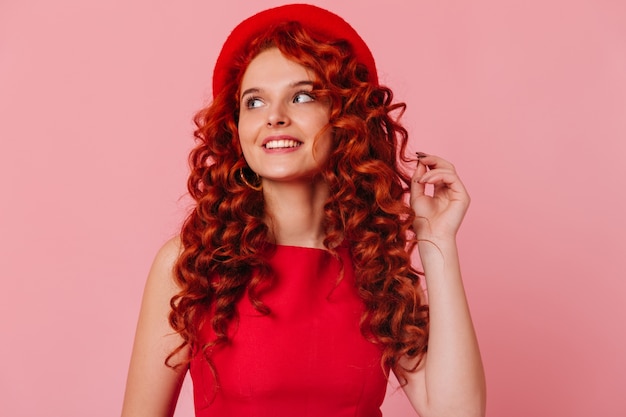 Foto gratuita encantadora chica pelirroja con sonrisa mira hacia otro lado. mujer con sombrero y top rojo toca el pelo.