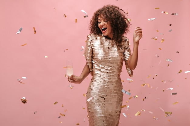 Encantadora chica con pelo corto ondulado en vestido beige brillante sosteniendo copa con champán y posando con confeti en pared rosa.