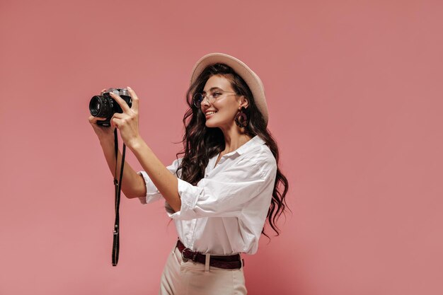 Encantadora chica de pelo largo ondulado con anteojos modernos blusa ancha clara pantalones beige con cinturón y sombrero sonriendo y posando con una cámara genial