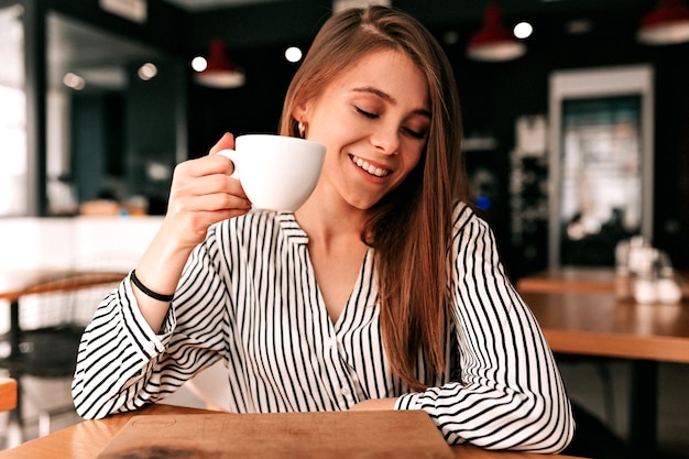 Foto gratuita encantadora chica de pelo largo sentada en la cafetería con café y tímidamente mira hacia otro lado