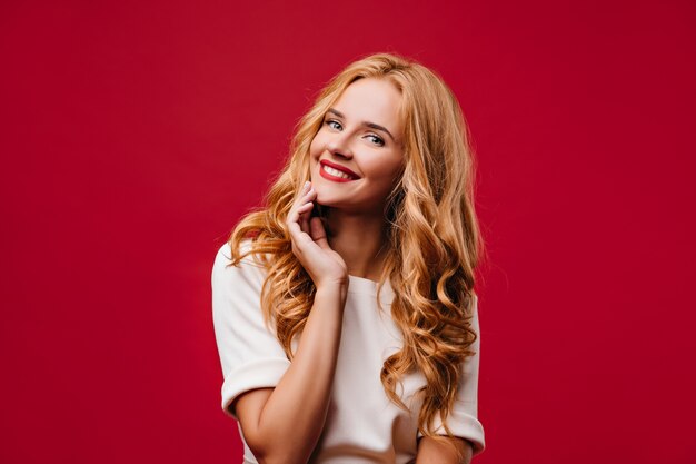 Encantadora chica de pelo largo sonriendo durante la sesión de fotos. Señora riendo alegre de pie en la pared roja.