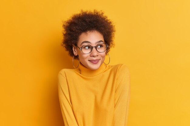 Encantadora chica de pelo rizado mira a un lado con expresión alegre, lleva gafas transprent de cuello alto casual admira algo hermoso se siente feliz.