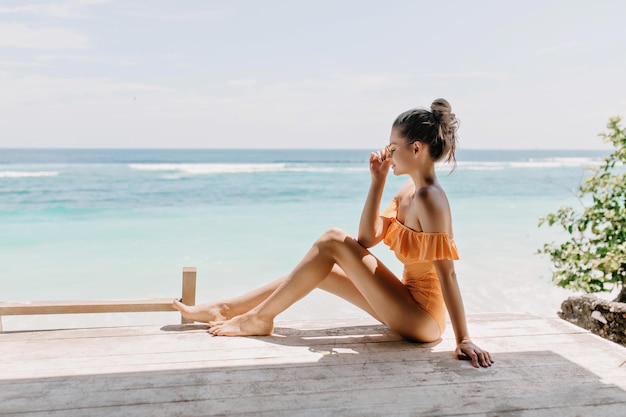 Encantadora chica con piel bronceada relajándose en una isla exótica por la mañana Foto al aire libre de una joven despreocupada en traje de baño naranja relajándose en la costa del océano