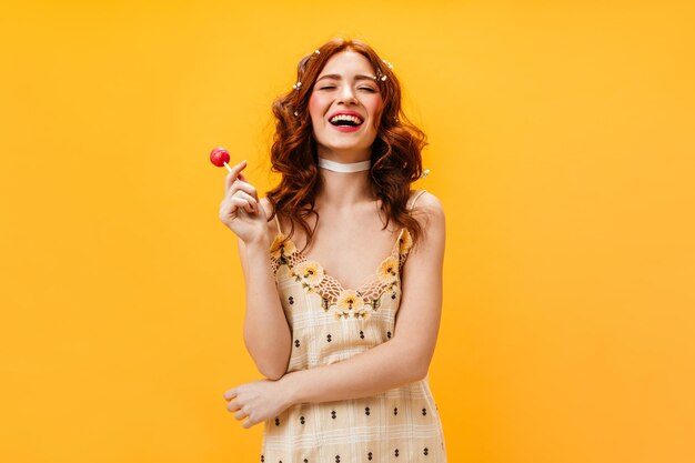 Encantadora chica positiva con vestido amarillo sostiene piruleta y se ríe con fondo naranja