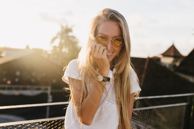 Foto gratuita encantadora chica en reloj de pulsera posando con sonrisa tímida sobre fondo de cielo.